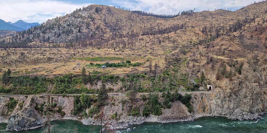 The rugged, rocky brown peaks of Canada are spotted with trees and sits next to a rushing river.  